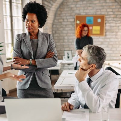 Female African American entrepreneur and her colleagues communicating on a business meeting in modern office. There are people in the background.